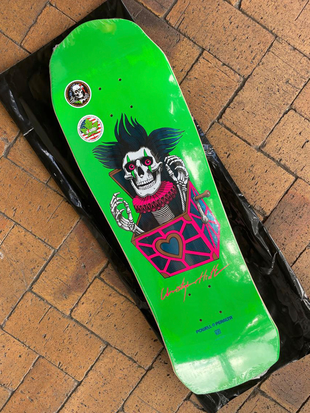 A bright green skateboard deck featuring a graphic of a skeleton in a box with pink and black details, photographed on a black plastic sheet over a brown brick floor.