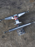 Two silver metal skateboard trucks with red and white bearings, displayed on a textured gray concrete surface.