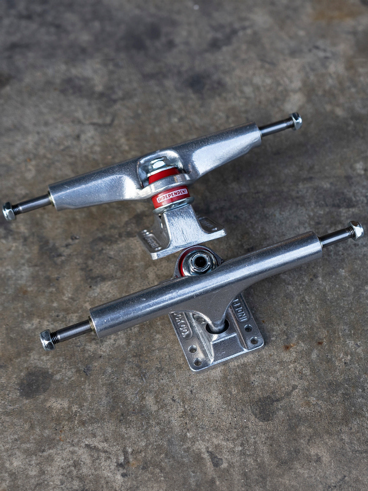 Two silver metal skateboard trucks with red and white bearings, displayed on a textured gray concrete surface.