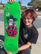 A person outdoors holding up a bright green Powell Pissarro skateboard with a skull jack-o'-lantern graphic. The person is wearing a black t-shirt and silver jewelry, with a skatepark and trees visible in the background.