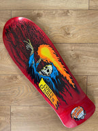 A Santa Cruz skateboard deck featuring a red background with a grim reaper graphic, placed diagonally on a light brown wooden floor. The graphic includes a skeleton in a blue robe with a flaming head, and the Santa Cruz logo is visible in the bottom right corner.