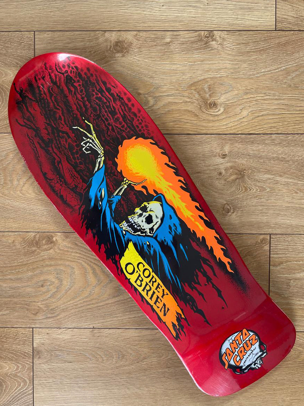 A Santa Cruz skateboard deck featuring a red background with a grim reaper graphic, placed diagonally on a light brown wooden floor. The graphic includes a skeleton in a blue robe with a flaming head, and the Santa Cruz logo is visible in the bottom right corner.
