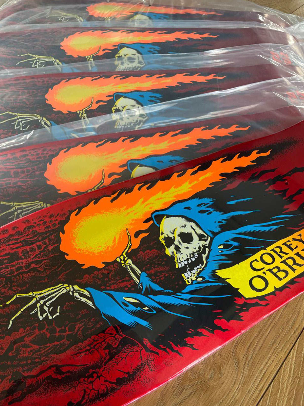 A close-up of a skateboard deck wrapped in clear plastic, showing a graphic of a hooded skeleton holding a flaming ball of fire. The deck has a red background with black, blue, orange, and yellow artwork, and the text 'COREY O'BRIAN' is visible on the right side.