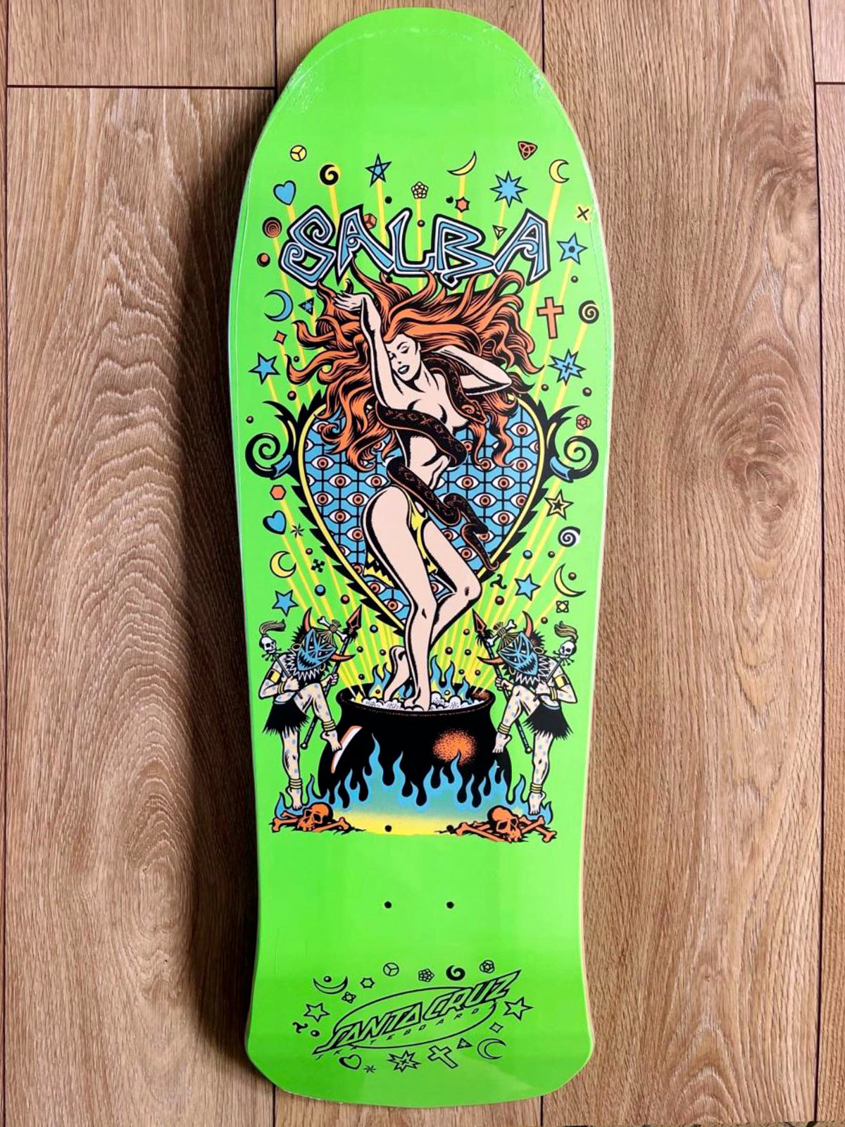 A bright lime green Salva skateboard deck with a detailed psychedelic graphic of a woman in a bikini standing on a cauldron, surrounded by stars, moons, and the word 'Salva' in stylized lettering, photographed flat on a wooden floor.