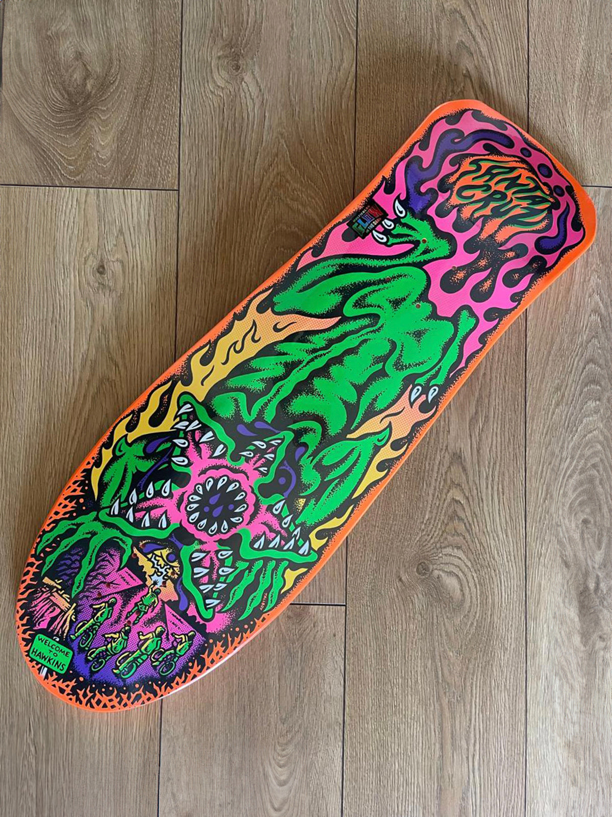 A skateboard deck with a vibrant, psychedelic monster graphic in neon green, pink, purple, yellow, and orange, photographed diagonally on a light brown wooden floor.