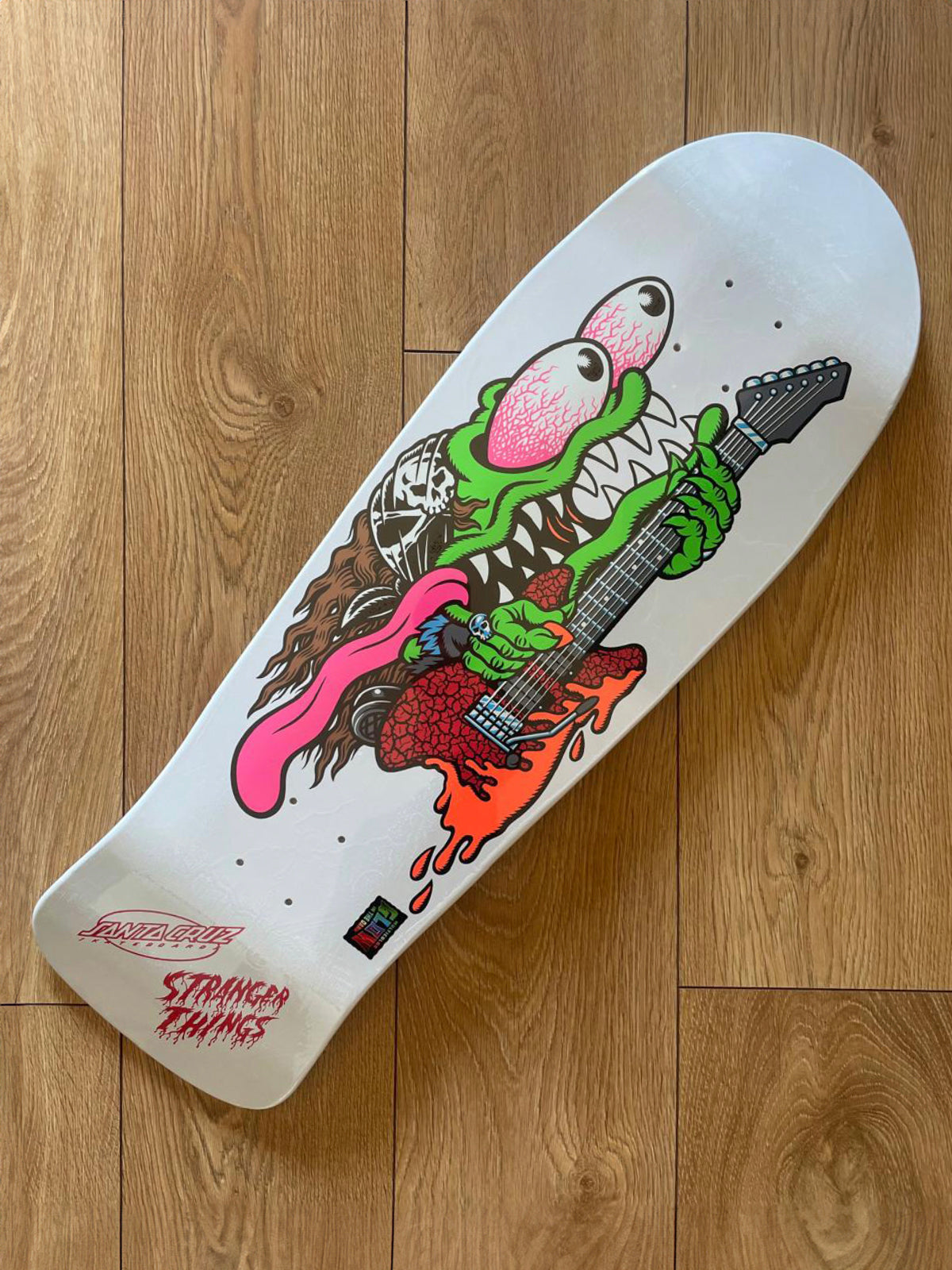 A white skateboard deck featuring a colorful graphic of a green monster playing a guitar, with the text 'STRANGLER THINGS' in red at the tail, photographed on a wooden floor.