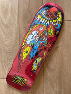 A Santa Cruz skateboard deck with a vibrant red base and a detailed comic-style graphic featuring a character in blue, yellow flames, and a red skull, photographed on a light wood floor.