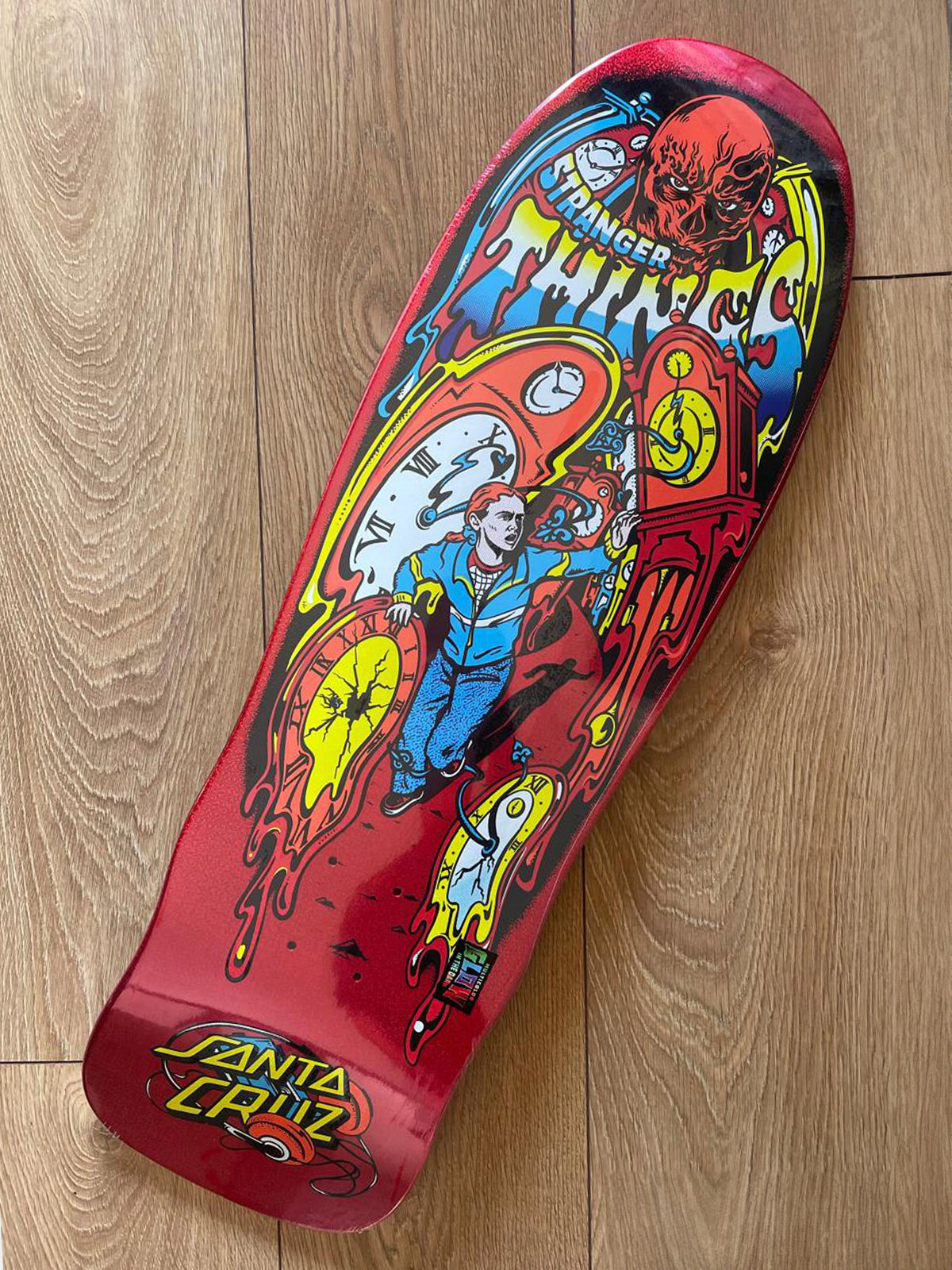 A Santa Cruz skateboard deck with a vibrant red base and a detailed comic-style graphic featuring a character in blue, yellow flames, and a red skull, photographed on a light wood floor.
