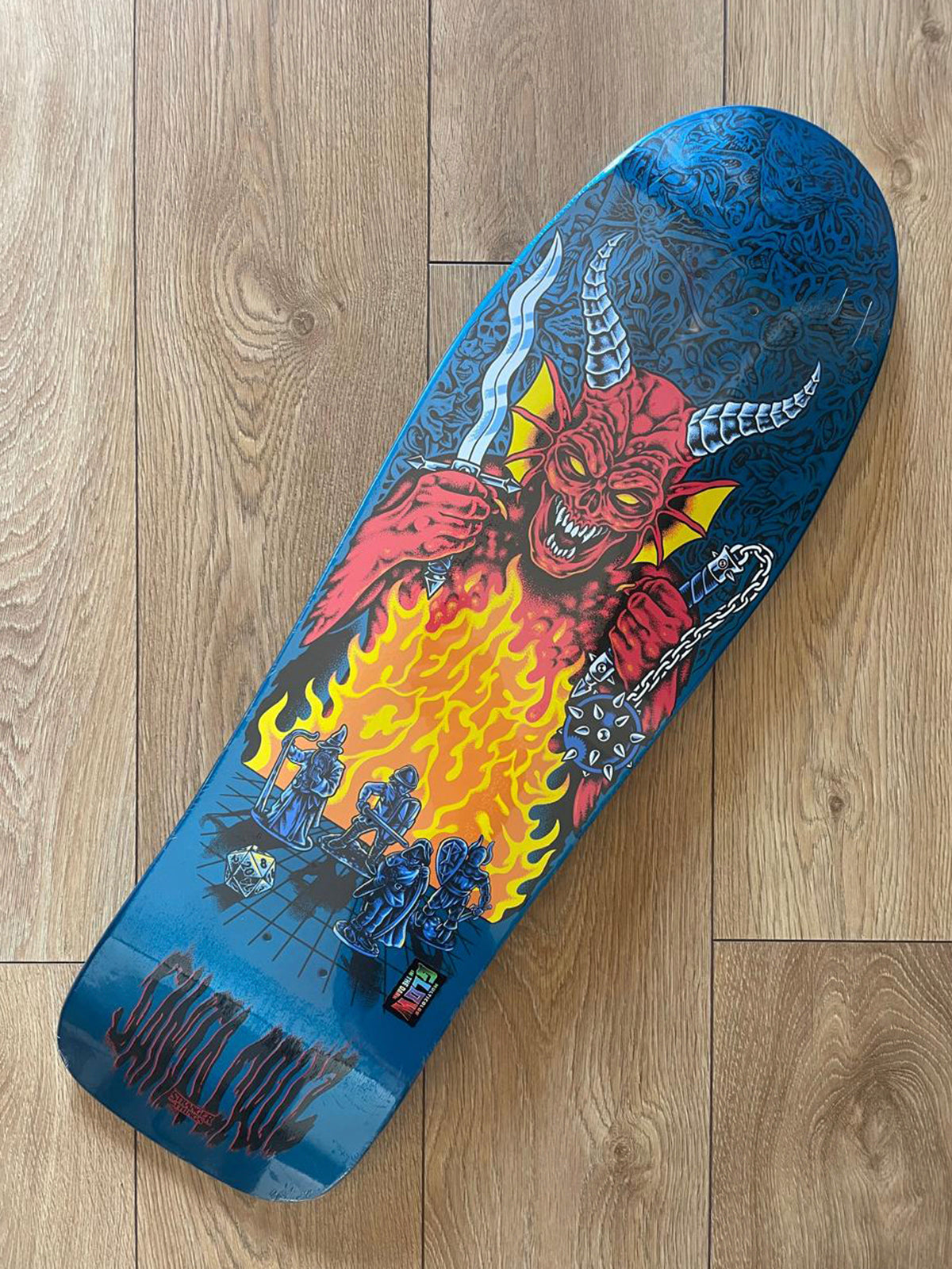 A skateboard deck with a detailed graphic of a red devil holding a sword and chains, with yellow flames and blue machinery on a wood plank floor.