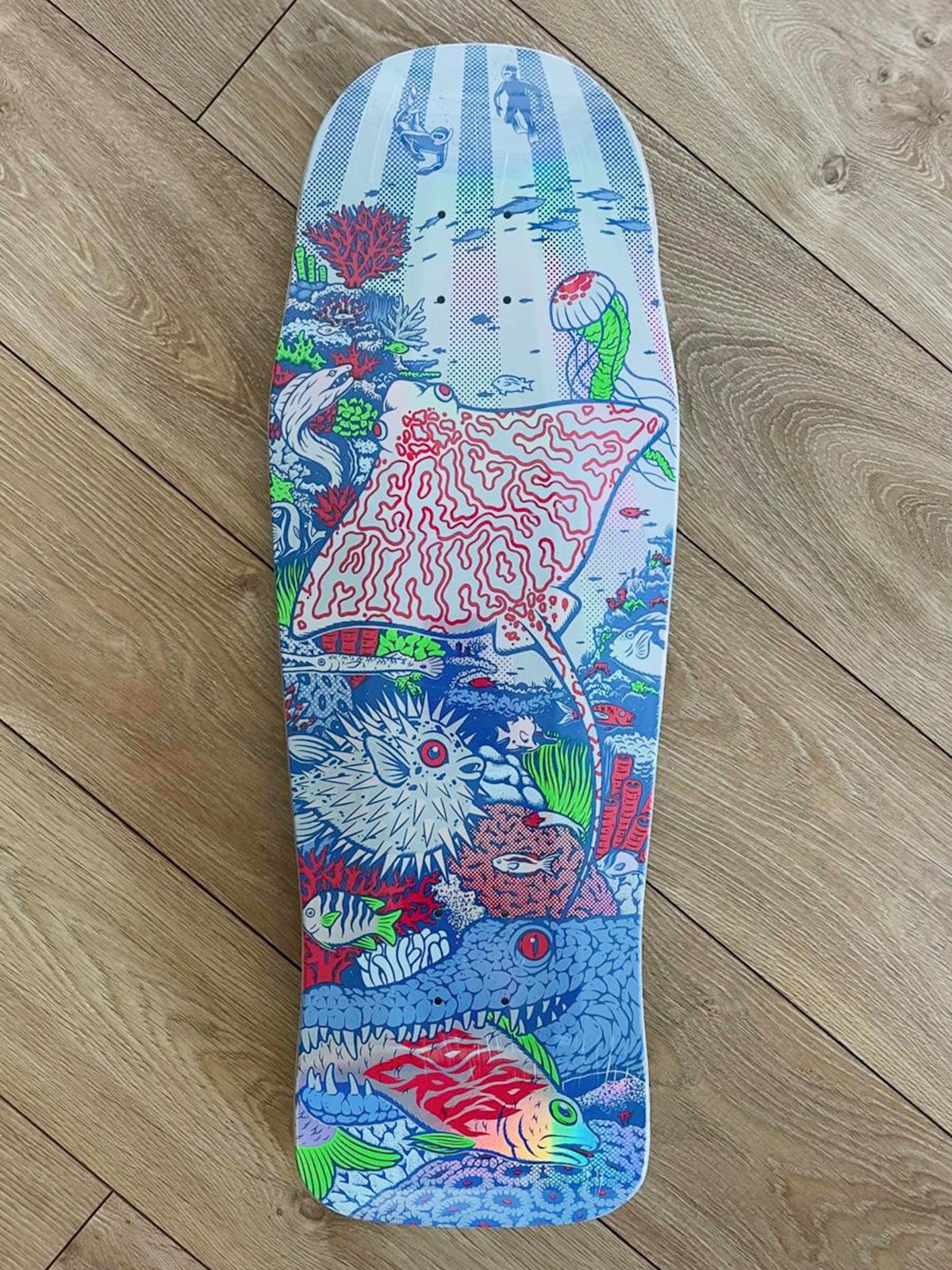 A skateboard deck with a highly detailed, colorful underwater illustration featuring a stingray, sea urchins, coral, and a red fish, photographed flat on a light brown wooden floor.